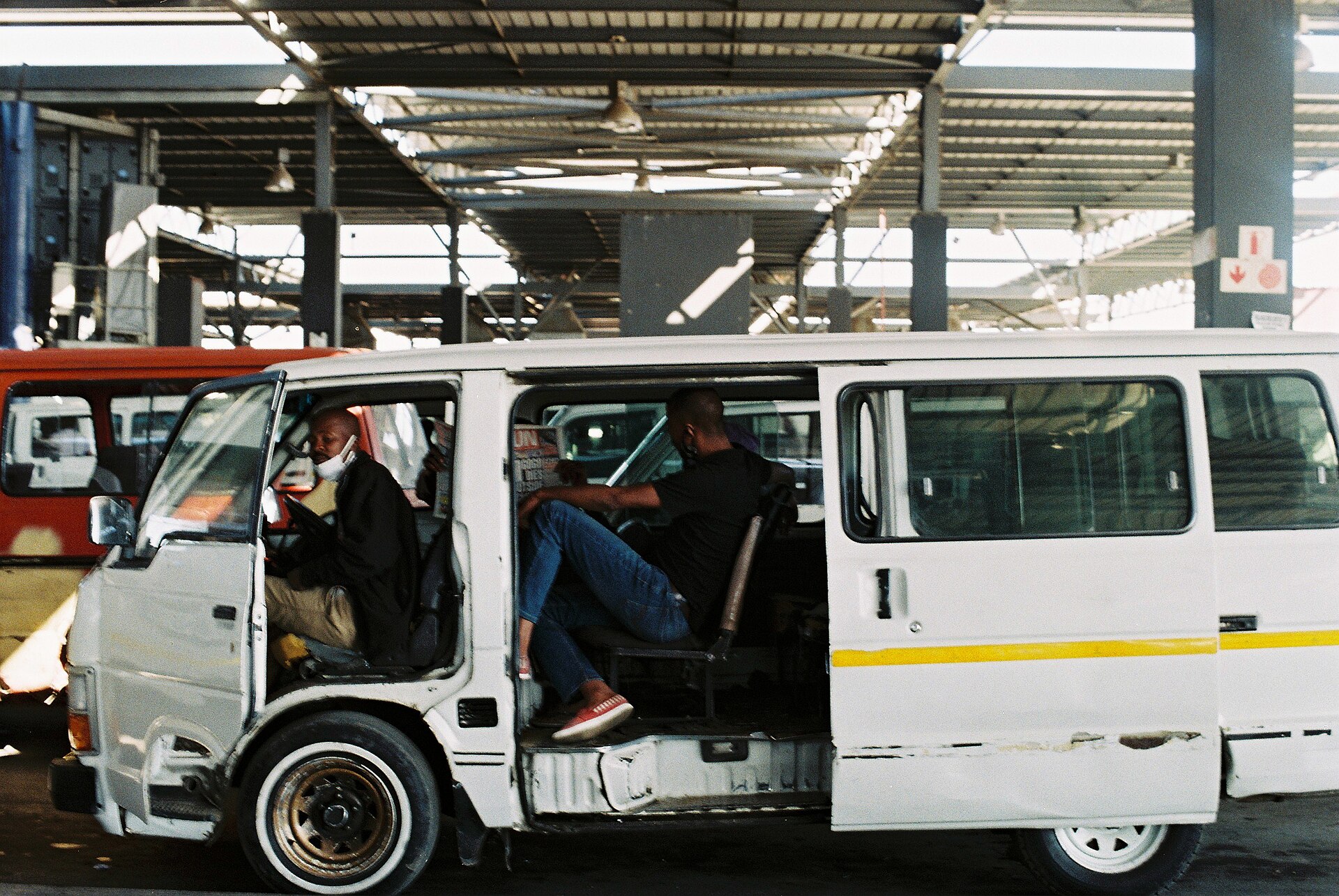 Two men sitting inside a white minibus at a depot, one reading a newspaper and one using a phone.